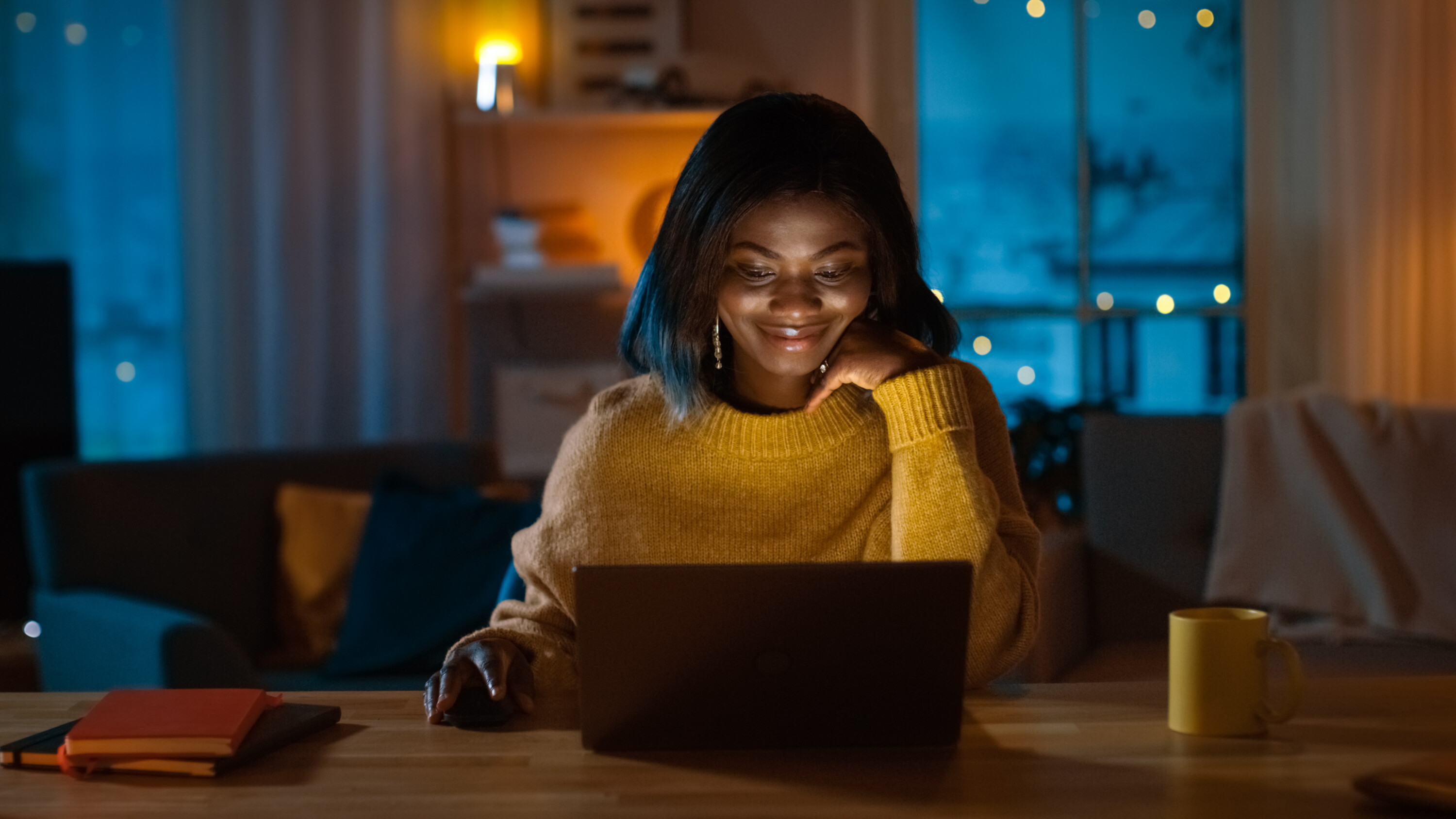 Portrait of Girl Uses Computer while Sitting at Her Desk at Home, She's Wearing Warm Sweater. In the Evening Creative Woman Works on a Computer In Her Cozy Living Room.